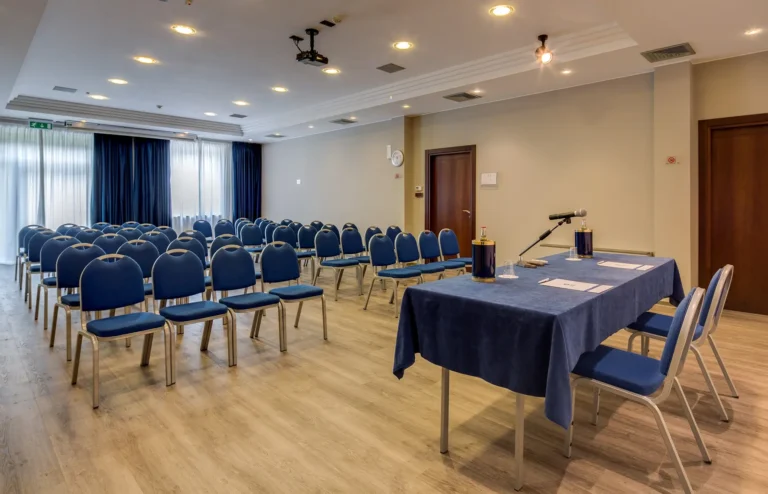 Sala meeting con sedie disposte a teatro, pavimento in parquet, grandi vetrate sul fondo, porte d'ingresso a destra e tavolo relatori in primo piano