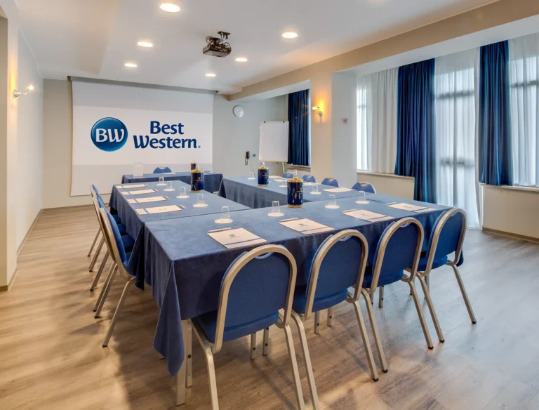 Sala meeting con disposizione a ferro di cavallo, tavolo relatori sul fondo, pavimento in parquet e schermo con logo Best Western proiettato