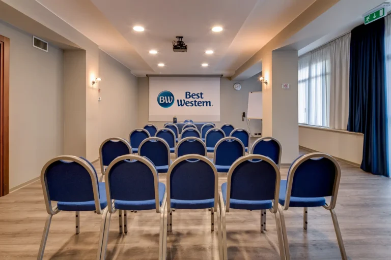 Sala riunioni con sedie disposte a teatro, pavimento in parquet, grande finestra sul lato destro e schermo per proiettore sul fondo con logo Best Western