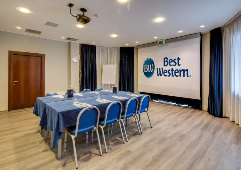 Sala riunione con allestimento a tavolo unico centrale, schermo per proiettore sul fondo, finestra sulla destra e porta d'ingresso a sinistra