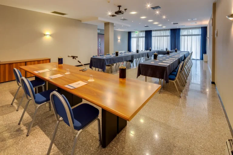 Ampia sala meeting, illuminata da tre grandi porte-finestre sul fondo, con tende bianche e tende oscuranti blu. Le pareti della sala sono beige e sul lato sinistro è presente una colonna. In primo piano c'è il tavolo dei relatori, in legno naturale con tre sedie blu. La sala è allestita con tavoli disposti a ferro di cavallo, ricoperti da tovaglie blu e circondati da sedie blu. La sala può ospitare 30 persone.