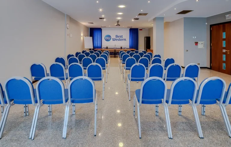 Grande sala riunioni con pavimenti in marmo e schermo per proiettore sul fondo, con logo Best Western. Il tavolo dei relatori si trova sul fondo della sala, le sedie del pubblico sono in primo piano e disposte a teatro. Sono presenti 48 sedie, in metallo grigio e tessuto blu. Sul lato destro della sala sono presenti una colonna e la porta d'ingresso, in legno e vetro.